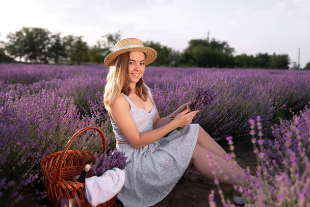 A young beautiful girl sitting among the lavender bushes holding a bouquet of flowers in the summer at sunset. field of blooming lavender and a lady in a hat. Nature concept. camping - Fotó, kép