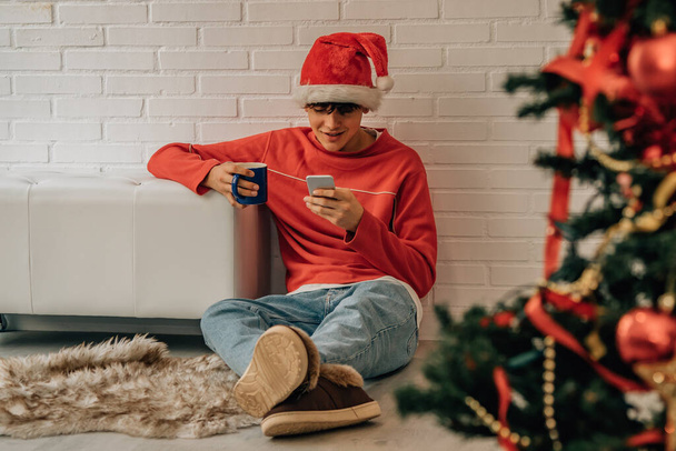 adolescent garçon avec téléphone mobile et santa claus chapeau à la maison pour Noël - Photo, image