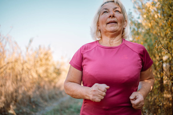 Портрет счастливой активной красивой пожилой женщины, бегущей утром на свежем воздухе в осеннем лесу. Спорт, кардио, концепция тренировок. Бег на свежем воздухе. - Фото, изображение