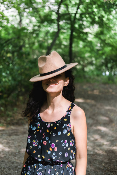Young millennial woman in dress and beige hat stands in forest happy and relaxed, enjoys nature and tranquility. Travel alone concept. - Фото, изображение