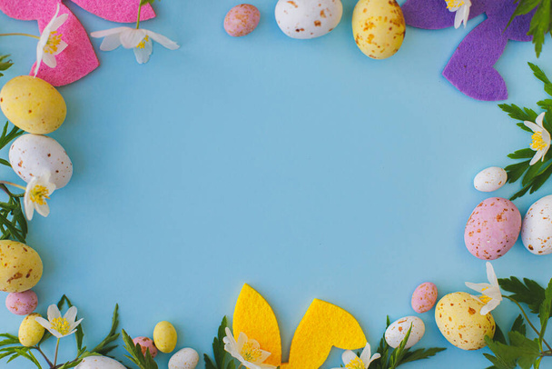 Colorful Easter bunnies, chocolate eggs and spring flowers frame on blue background, flat lay with space for text. Happy Easter! Pink and yellow artificial decor and fresh flowers. Easter hunt - Photo, Image