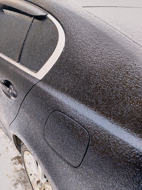 Ένα μαύρο επιβατικό αυτοκίνητο μετά από παγωμένη βροχή το χειμώνα. Το αυτοκίνητο καλύπτεται με ένα παχύ στρώμα από διαφανές γυαλιστερό πάγο. Ένα αυτοκίνητο σε κακές καιρικές συνθήκες το χειμώνα. - Φωτογραφία, εικόνα