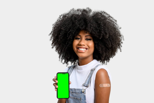 fille noire sans écran facial tient le téléphone portable près de son bras avec l'autocollant de vaccin, entièrement immunisé, clé chromatique sur l'écran du téléphone portable, sur fond gris - Photo, image