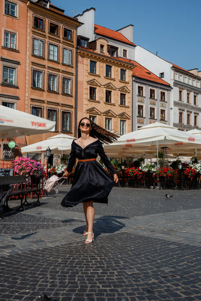 幸せな、笑顔の女性がスタイリッシュな黒のドレスとサングラスを着てワルシャワの街を歩く。ワルシャワの朝有名な広場で鳩を分散実行している女性. - 写真・画像