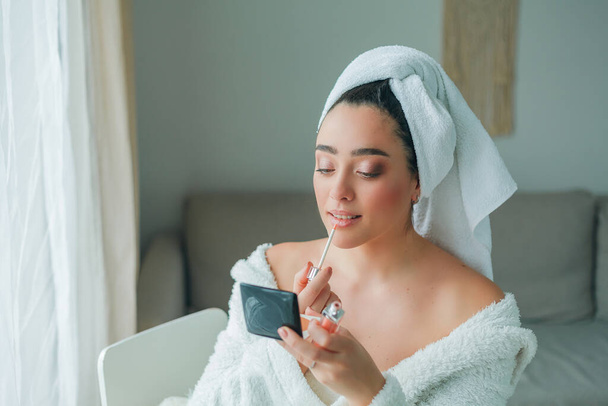 Young beautiful woman in terry white dressing gown makes herself beautiful makeup at home. Model holds makeup brush and cosmetics in her hands. Cozy home. - Φωτογραφία, εικόνα
