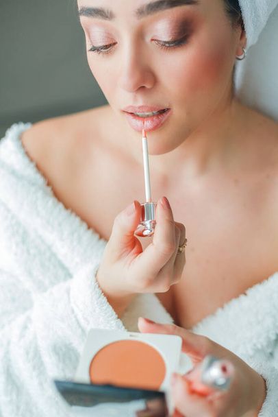 Young beautiful woman in terry white dressing gown makes herself beautiful makeup at home. Model holds makeup brush and cosmetics in her hands. Cozy home. - Foto, imagen