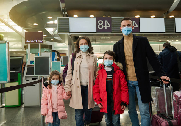Portrait of a young family wearing protective medical masks traveling during a pandemic, standing with luggage and suitcase at the airport while waiting to check in for a flight. Air travel concept - Fotografie, Obrázek