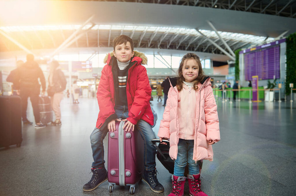 Happy beautiful Caucasian children in warm clothes, a handsome boy sits on a suitcase next to his younger sister- a cute little girl, at the international airport. Family airplane travel concept - Φωτογραφία, εικόνα