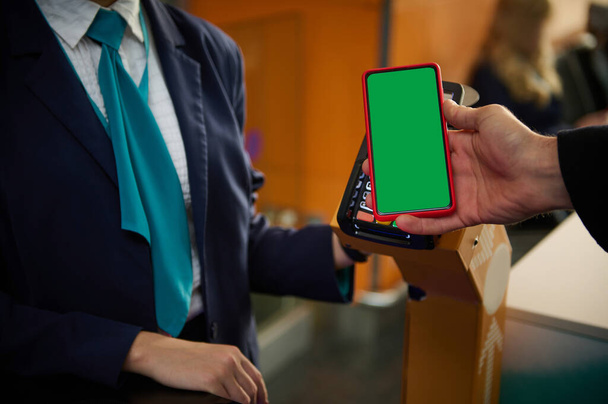 Close-up of man's hand holding mobile phone with green chroma key blank screen with copy space for mobile apps and advertising, near bank terminal while going through flight check-in board at airport - 写真・画像