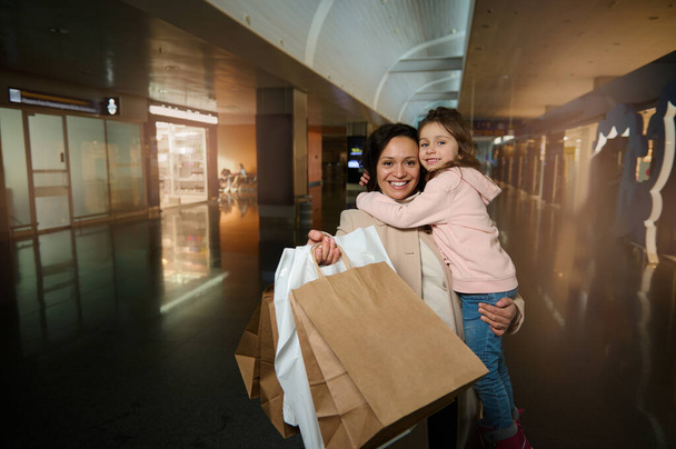 Happy mother holding and hugging her cute daughter in arms, smiling, looking at camera showing shopping bags standing in duty free area of departure international airport terminal, waiting for flight - Photo, image