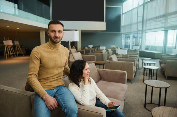 Attractive handsome Caucasian man looks at camera hugging his girlfriend, sitting in chair in an international airport lounge, waiting to board a flight. Multi ethnic couple travels on their honeymoon - Zdjęcie, obraz