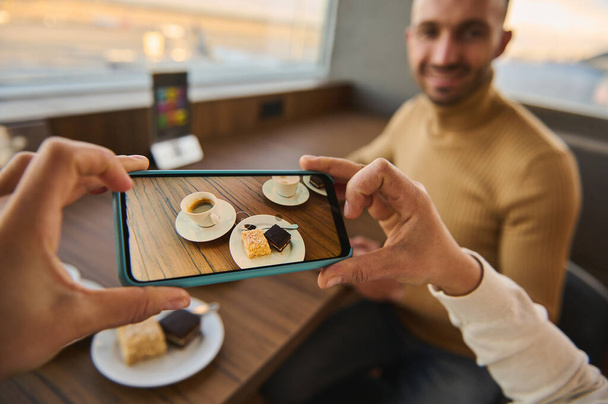 Woman hands taking photo on smartphone of cup of coffee and cakes on wooden table with blurred handsome man sitting at panoramic windows overlooking the planes on runways. Mobile phone live view mode - 写真・画像