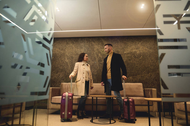 Handsome attractive businessman talking to his business partner, beautiful woman standing with suitcases at the exit from the conference room in the airport terminal ready to board the flight together - 写真・画像