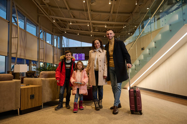 家族旅行を楽しんでいる幸せな子供や両親を喜ばせ、スケジュール付きのフライト情報パネルに対する空港出発ターミナルのラウンジゾーンにスーツケースと一緒に立っています。パンデミック後の旅 - 写真・画像