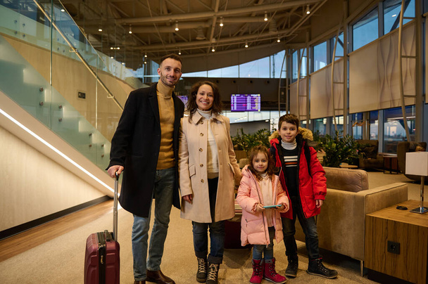 Happy family enjoying the trip together, stands with luggage in the lobby of the airport departure terminal, waiting for boarding, on the background of the information panel with the flight schedule - Foto, Imagen