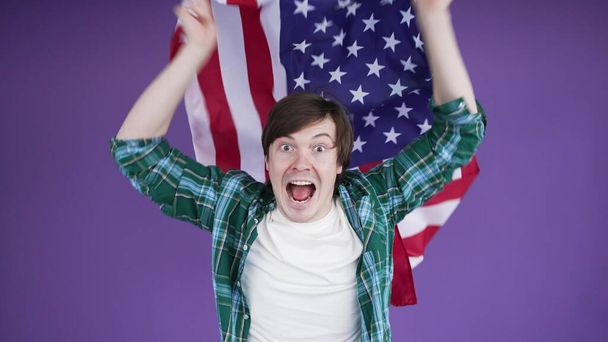 Young emotional man waving american flag and shouting on purple background - Фото, изображение