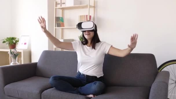 Mujer joven con gafas de realidad virtual jugando juegos virtuales en el sofá  - Imágenes, Vídeo
