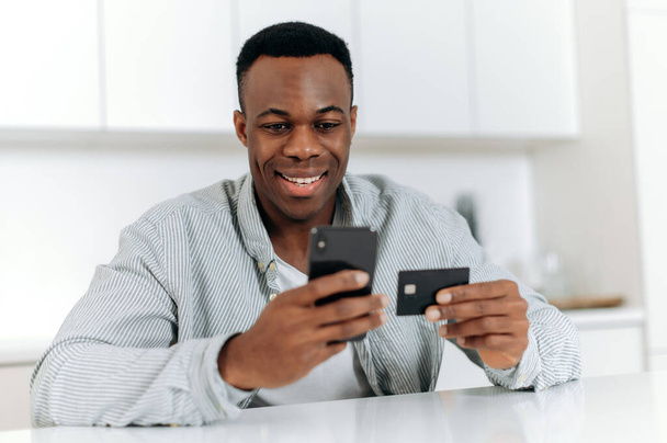 正のアフリカ系アメリカ人の若者が自宅でキッチンに座って、カジュアルな服装で、オンラインショッピングのためのスマートフォンやクレジットカードを使用して、支払い、オンライン取引、笑顔のためのデータを入力しています - 写真・画像