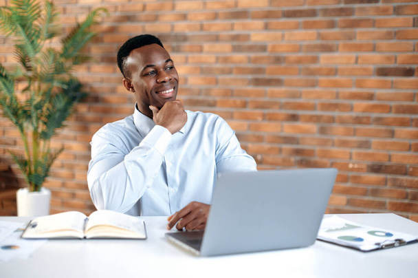 Proud successful African American business man, top manager, head of department, sits at the workplace in modern office, looks thoughtfully to the side, smiles, ponders a project, dreams of a vacation - Фото, зображення