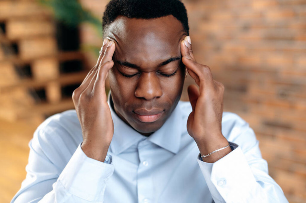 Close-up of an exhausted african american man experiencing headache, migraine, overwork from working at the computer, sad guy is sick, needs treatment and rest, closed his eyes, massages temples - Fotografie, Obrázek