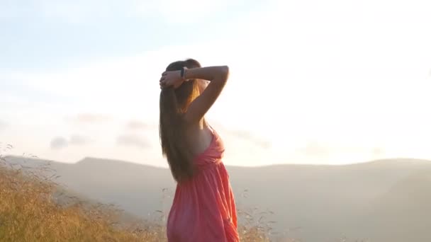 Jovem mulher feliz viajante em vestido vermelho em pé na encosta gramada em uma noite ventosa em montanhas de verão desfrutando de vista da natureza ao pôr do sol - Filmagem, Vídeo
