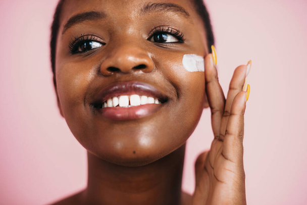 Close-up portrait of a black woman applying moisturizing cream on face. Body care. Isolated over pink backdrop - Foto, Imagen