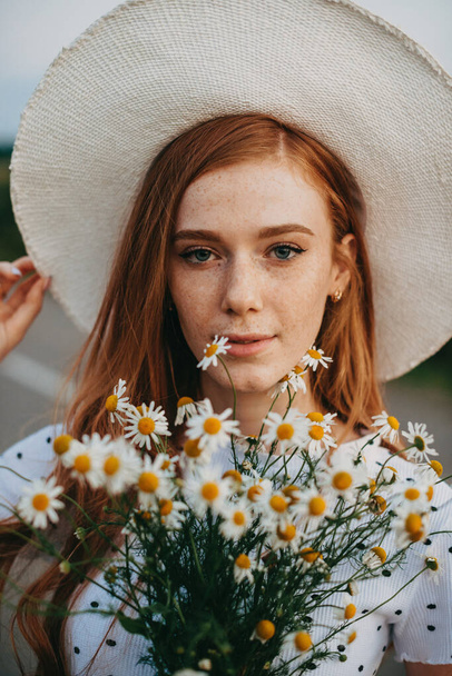 Şapkalı kızıl saçlı bir kadının yakın plan portresi ve güneşli bir yaz gününde elinde bir buket çiçek. Papatya tarlasında bir kadın. Doğal güzellik. Mutlu insanlar.. - Fotoğraf, Görsel