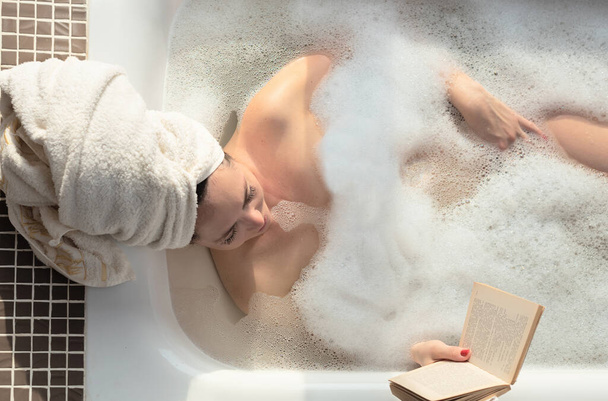 Young woman takes a bath with lush foam near the window reading the book. Personal hygiene, healthcare, relax at home concept - Photo, image