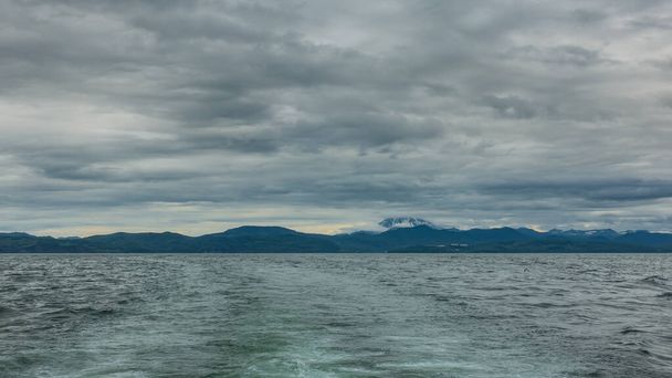 On the surface of the Pacific Ocean, ripples are visible, a foam trail from the yacht. A mountain range against a cloudy sky in the distance. A snow-covered volcano is hiding in the clouds. Kamchatka - Photo, Image