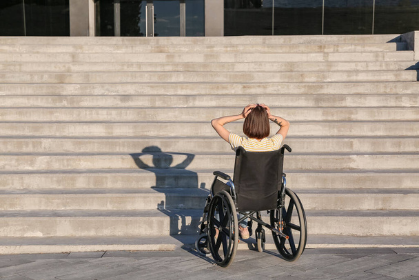 Αγχωμένη νεαρή γυναίκα σε αναπηρικό καροτσάκι κοντά σε σκάλες σε εξωτερικούς χώρους - Φωτογραφία, εικόνα
