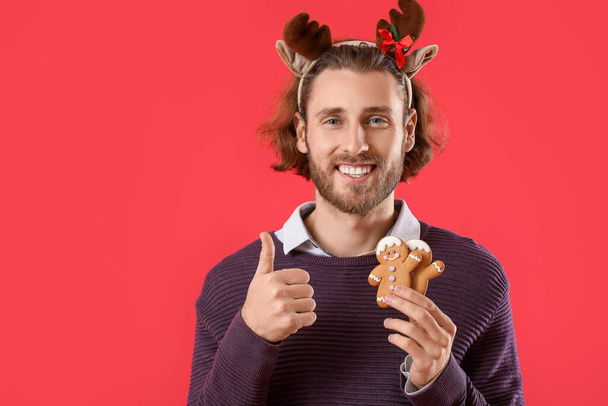 Handsome man in deer horns with gingerbread cookies showing thumb-up on red background - Foto, immagini