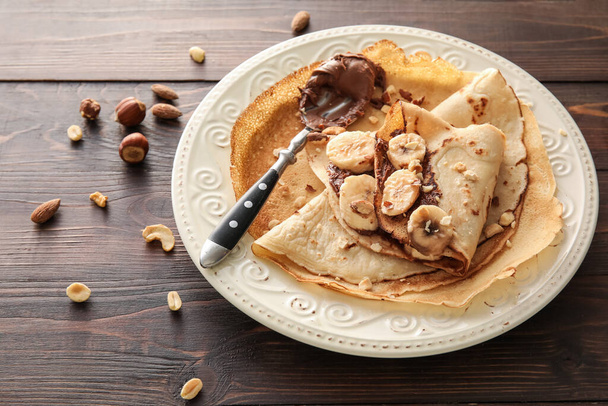 Plate of tasty thin pancakes with chocolate paste, cut banana and nuts on wooden background - Φωτογραφία, εικόνα