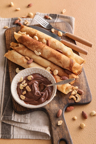 Wooden board of tasty thin pancakes with chocolate paste and nuts on beige background - Foto, immagini