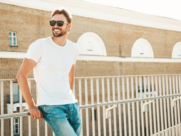 Portrait of handsome smiling  hipster lambersexual model.Stylish man dressed in white T-shirt and jeans. Fashion male posing on the street background near fence at the beach  - Foto, imagen
