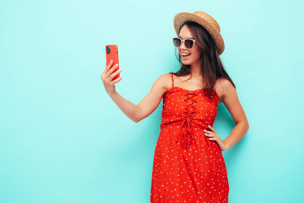 Young beautiful smiling female in trendy summer red dress. Sexy carefree woman posing near blue wall in studio. Positive brunette model having fun in hat and sunglasses. Taking selfie photos - Foto, imagen