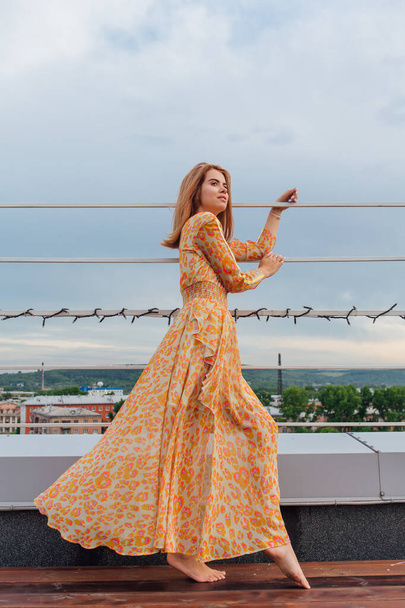 Beautiful young woman in long leopard dress standing with bare feet on the stairs on the top of the roof of a building. - Foto, imagen