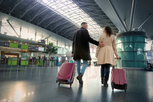 Longitud completa de dos personas guapas hombre y mujer, pareja joven con maletas caminando a lo largo de la sala de salida del aeropuerto internacional en el fondo borroso de los mostradores de facturación de vuelo - Foto, imagen