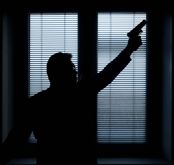 A man with a gun is standing by the window blind, ready to protect himself from another bad people, crime scene conceptual. silhouette of an armed man behind blinds - Fotografie, Obrázek