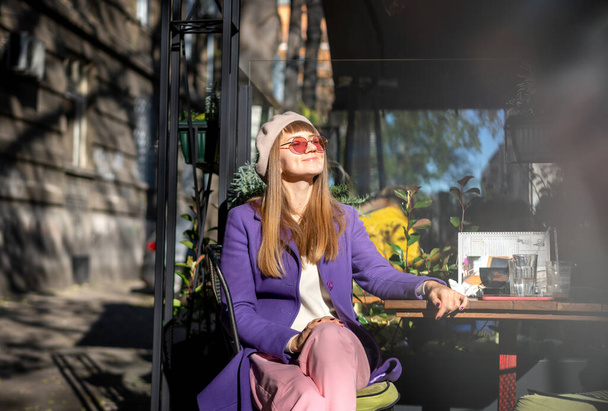Young cheerful woman in purple coat sitting in street cafe enjoying sunny spring day - Foto, afbeelding