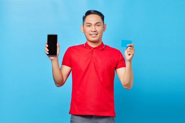 青い背景に隔離された空白の画面の携帯電話とクレジットカードを示す笑顔の若いアジア人男性の肖像画 - 写真・画像