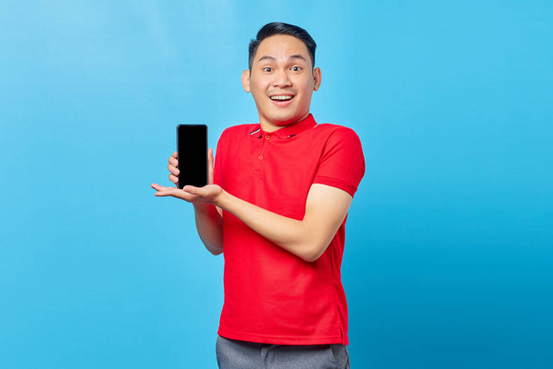 Portrait of cheerful young asian man in red shirt showing blank screen mobile phone and looking at camera isolated on blue background - Foto, Imagem
