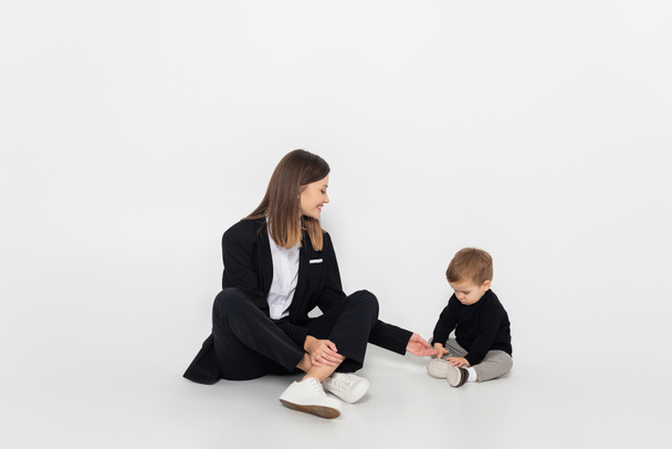 stylish and happy woman sitting near toddler son on grey - Foto, imagen