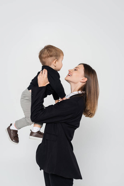 cheerful mother holding in arms toddler son isolated on grey - Foto, imagen