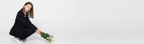 trendy young woman in black suit sitting and holding white flowers on grey, banner - Foto, afbeelding