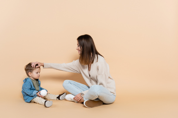 happy mother sitting and adjusting hair of toddler son on beige - Fotoğraf, Görsel