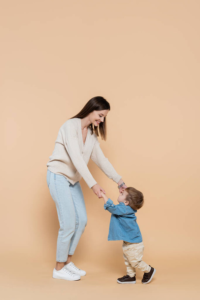 full length of cheerful mother standing and holding hands with toddler son on beige - Photo, Image