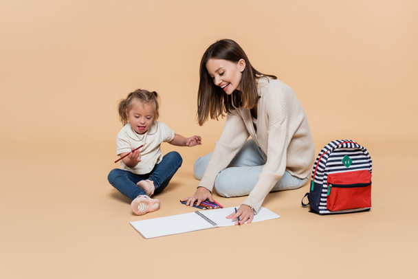 happy mother sitting near kid with down syndrome drawing near colorful pencils and backpack on beige - Photo, Image