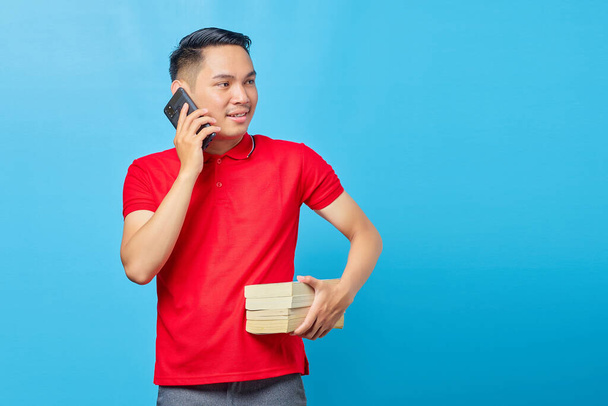 Muotokuva houkutteleva aasialainen mies puhuu älypuhelimella ja etsivät onnellinen pitäen kirja eristetty sinisellä pohjalla - Valokuva, kuva