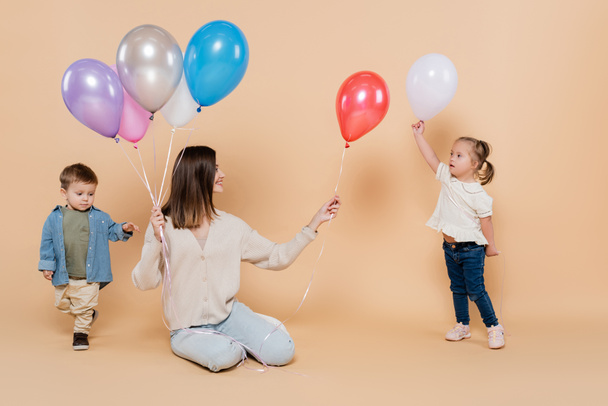 positive woman sitting near girl with down syndrome and toddler boy while holding colorful balloons on beige - Photo, Image