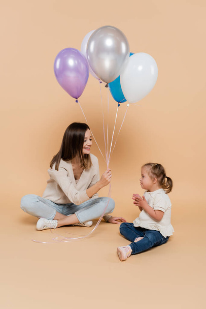 positive woman sitting near girl with down syndrome while holding colorful balloons on beige - Zdjęcie, obraz